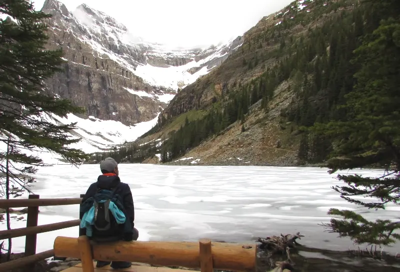 Persona sentada en un banco de madera contemplando el lago Lake Agnes helado frente a las montañas nevadas.