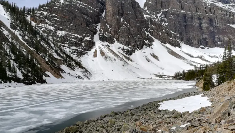 Vista desde el sendero lateral de Lake Agnes congelado, mostrando las montañas escarpadas y la orilla rocosa con nieve.