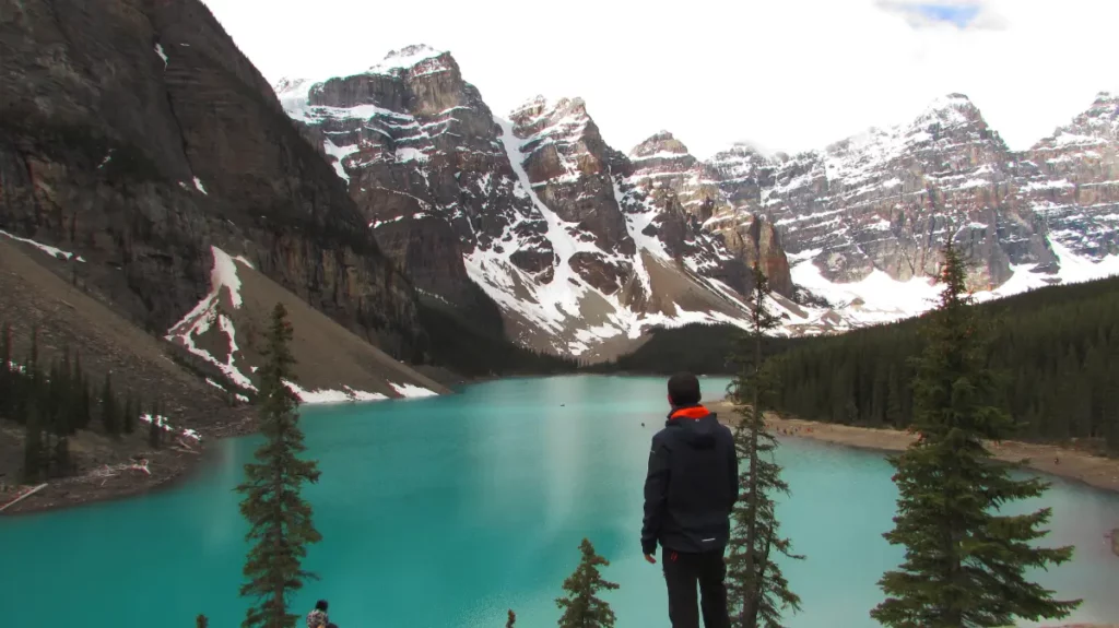 Hombre de espaldas contemplando la inmensidad del lago Moraine y las cumbres nevadas desde lo alto del mirador Rockpile.