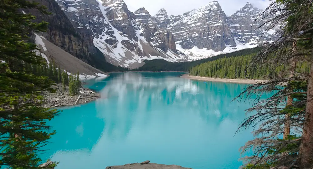 Panorámica del Lago Moraine enmarcada por abetos durante el ascenso al mirador Rockpile, con las montañas Ten Peaks reflejadas en el agua turquesa.