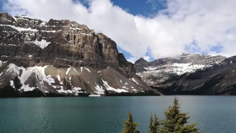 Vista panorámica de Bow Lake con sus aguas turquesas rodeado de imponentes montañas de roca y nieve bajo un cielo con nubes en Alberta, Canadá.
