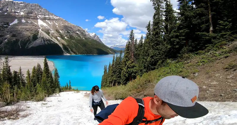 Dos personas ascendiendo por una lengua de nieve en la ladera de la montaña con el azul intenso de Peyto Lake al fondo.