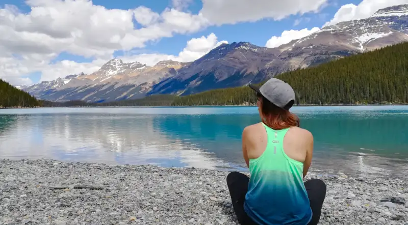 Persona de espaldas con camiseta degradada sentada en la orilla de piedras de Peyto Lake, disfrutando de las vistas al agua turquesa y las montañas.