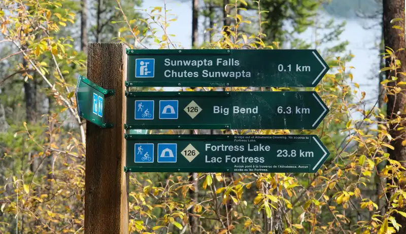 Cartel de madera con indicaciones verdes hacia Sunwapta Falls a 0,1 km, Big Bend y Fortress Lake en un sendero rodeado de hojas otoñales.